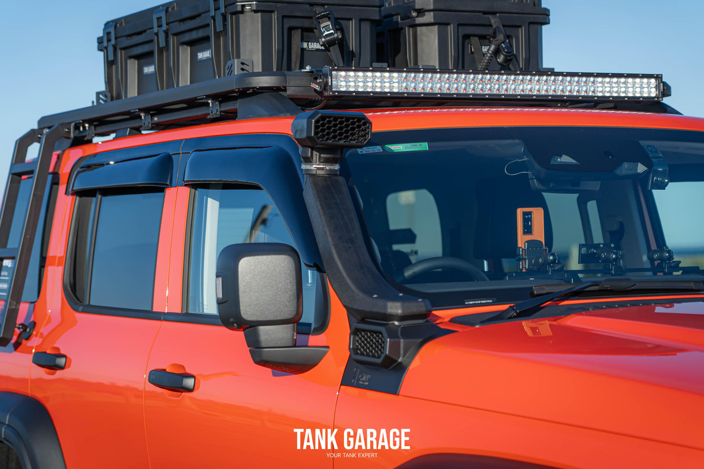 An orange Tank vehicle equipped with transparent Car Window Weather Shields. These shields are designed to provide protection from rain, wind, and debris while maintaining visibility and enhancing the vehicle's sleek look.- Tank Garage